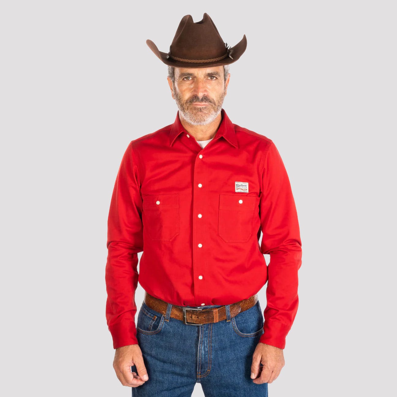 Man wearing a red shirt, blue jeans, and a brown cowboy hat on a gray background